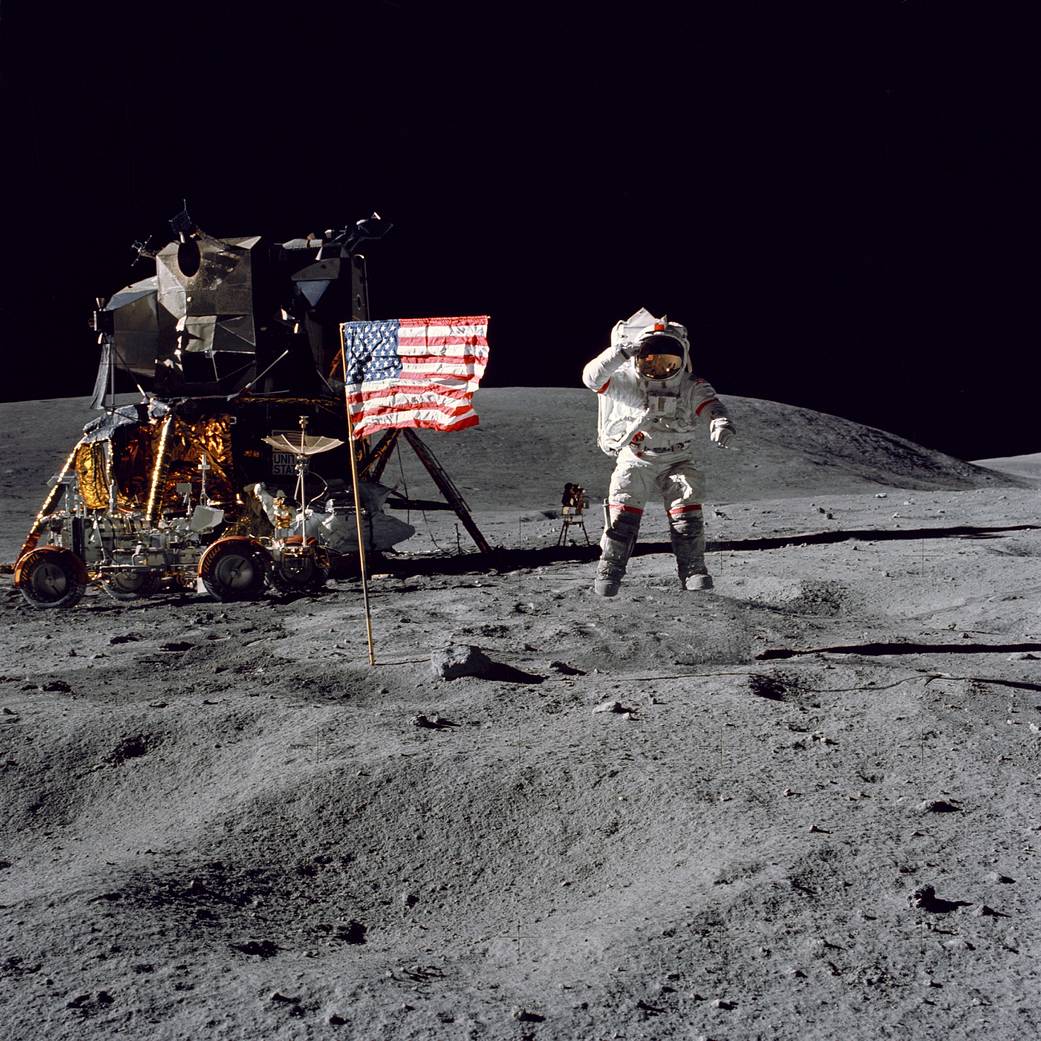 Un astronaute d'Apollo 16 saluant à côté du drapeau Américain, en arrière plan le module et le rover lunaires